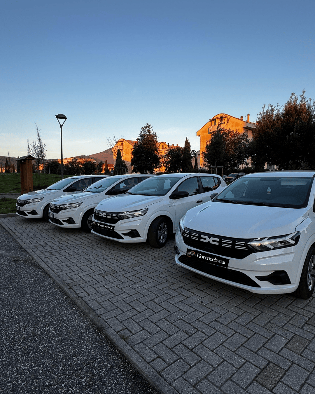 Fleet of new white Dacia rental cars parked in a row at Florence by Car headquarters, ready for customer pick-up under a clear evening sky.